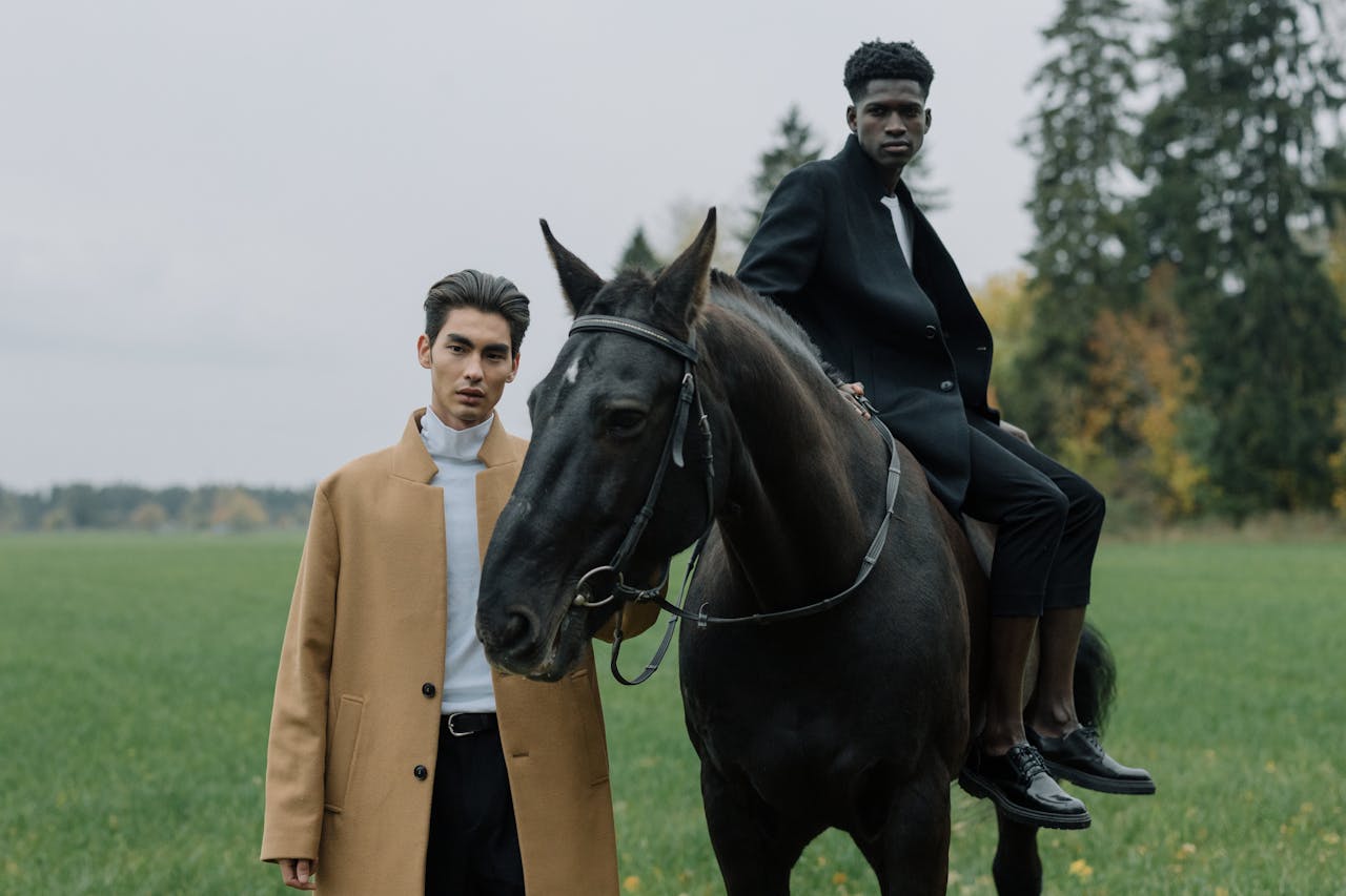 Two stylish models in elegant coats and a horse, set in a serene field.