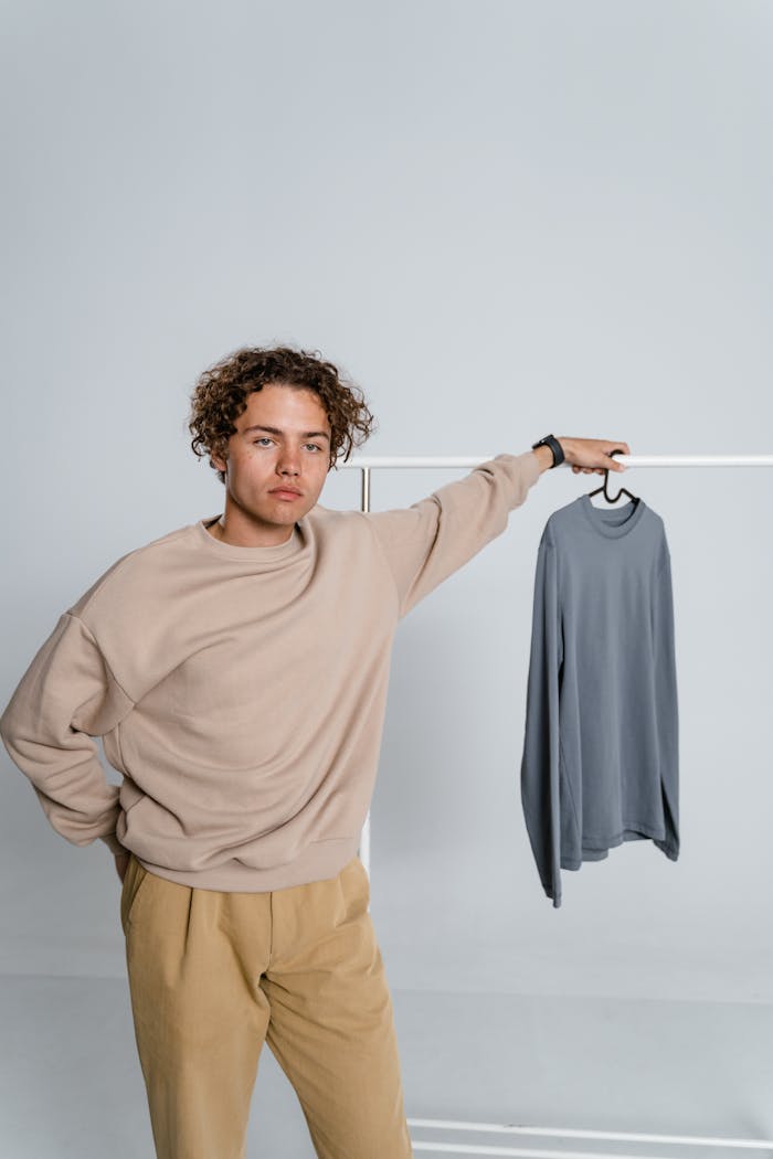 Young man with curly hair posing in a minimalist studio with a sweater on a hanger.